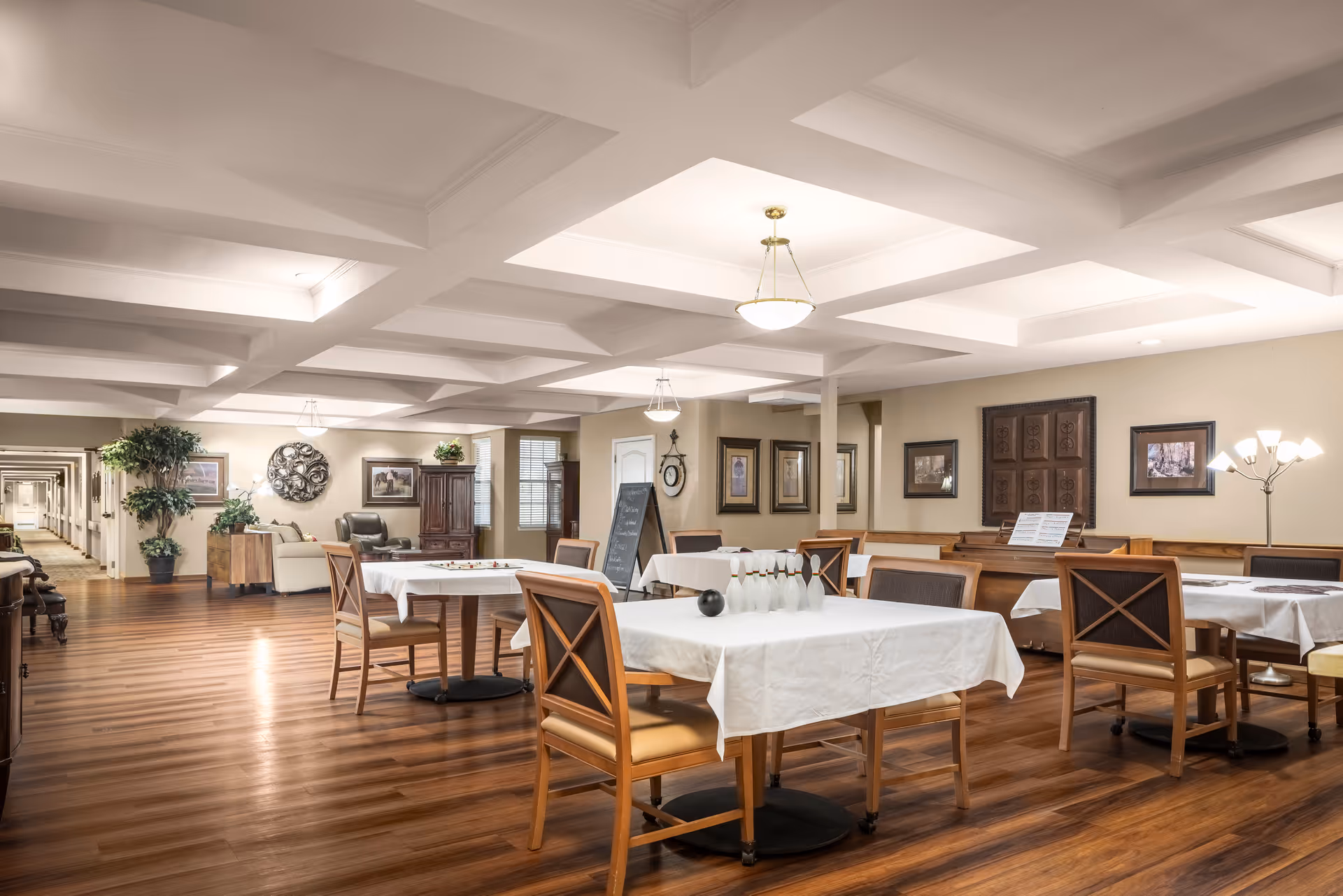 Spacious, well-lit dining/activity room with tables covered in white tablecloths, wooden chairs, and hardwood floors.