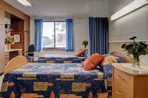 A shared bedroom in a senior living facility with two single beds covered in patterned bedspreads. Each bed has pillows and is positioned against opposite walls. There is a window with blue curtains letting in natural light, a wooden nightstand with a vase of flowers, and a wooden chair near the window. The room has a cozy and clean appearance.