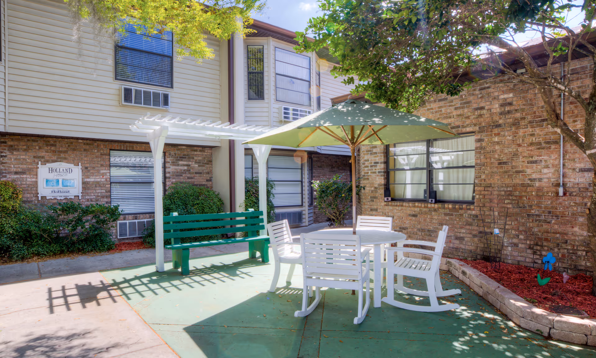 Outdoor patio area at Brookdale New Port Richey with a round white table, four white chairs, a green umbrella, a green bench, and surrounding brick and siding building walls with windows and greenery.