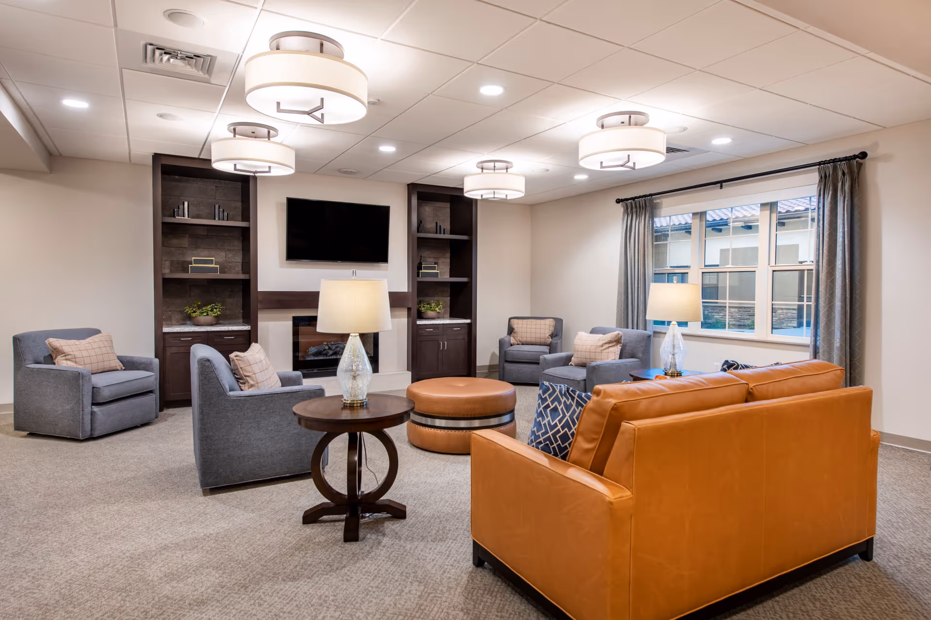 A cozy living room with a tan leather sofa, three gray armchairs with pillows, a round ottoman, two side tables with lamps, built-in dark wood shelves, a wall-mounted TV above a fireplace, and a window with gray curtains letting in natural light.