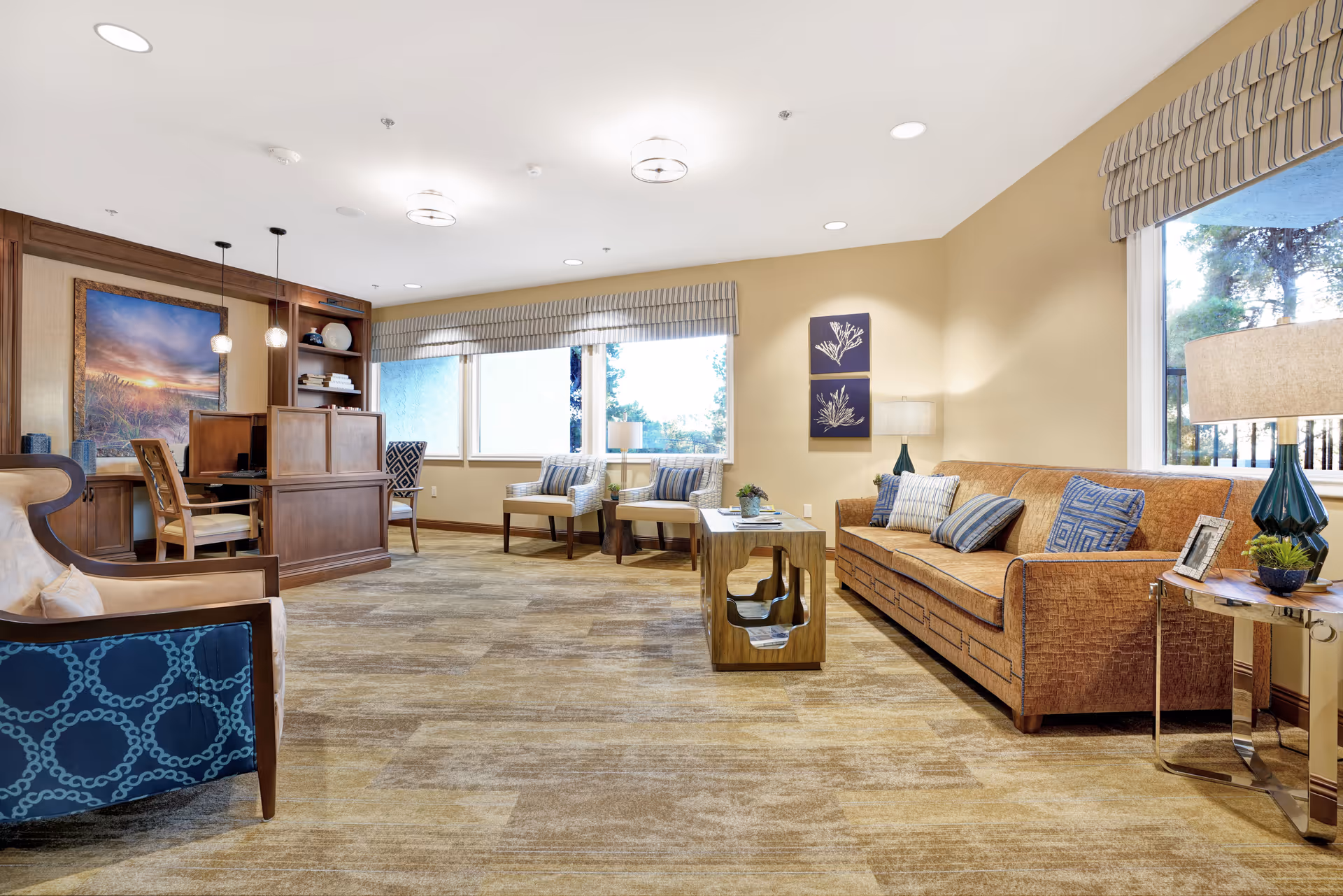 A bright and spacious living room area in a senior living facility with a brown sofa adorned with blue and white patterned pillows, two armchairs with striped cushions near large windows, a wooden coffee table with magazines, and a wooden desk with chairs and decorative items. The room has beige walls, carpeted flooring, and ceiling lights.