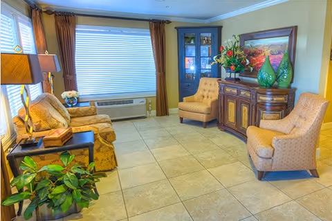 A cozy living room with beige tiled floor, a patterned sofa, two upholstered armchairs, a wooden side table with a lamp, a large window with brown curtains and blinds, a wooden cabinet with glass doors, a sideboard decorated with green vases and a floral arrangement, and a colorful framed painting on the wall.