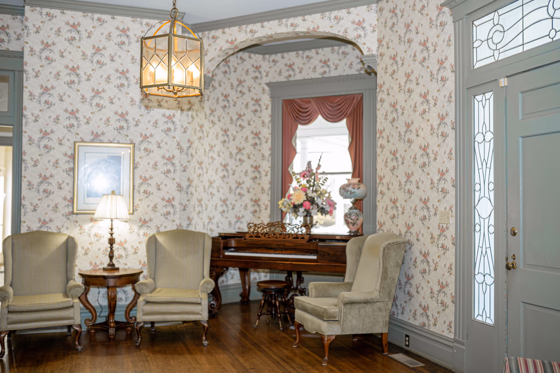 A cozy living room area with floral wallpaper, three upholstered armchairs, a wooden side table with a lamp, and a vintage wooden piano adorned with a floral arrangement and decorative vases. The room features a large window with red drapes and a door with decorative glass panels.