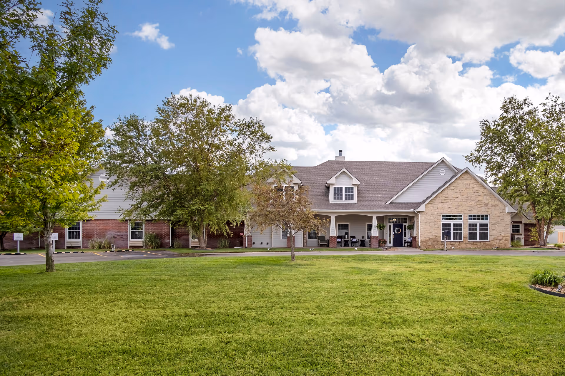 Front exterior view of a single-story building with a mix of brick and stone facade, surrounded by green grass and trees under a partly cloudy sky.