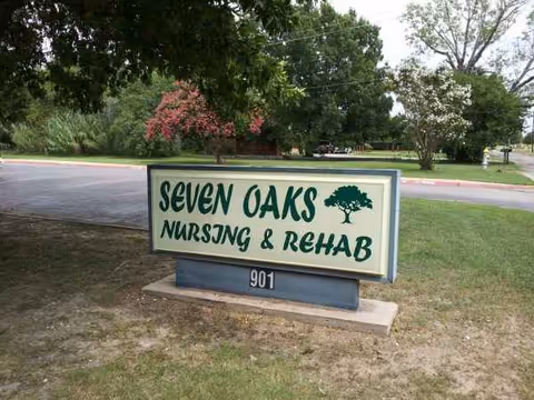 Outdoor sign for Seven Oaks Nursing & Rehab on grassy area with trees and road.