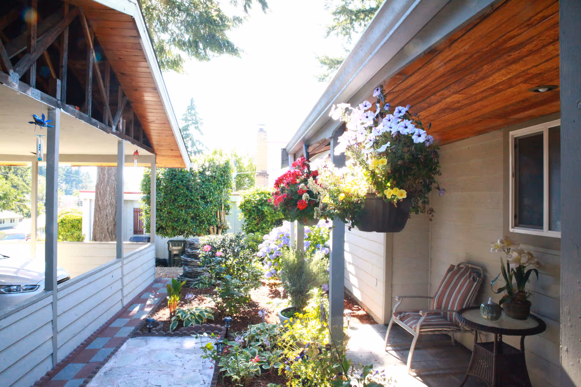 A bright outdoor garden area between two buildings with hanging flower pots, various plants, a small table with a potted plant, and a striped cushioned chair under a wooden ceiling.
