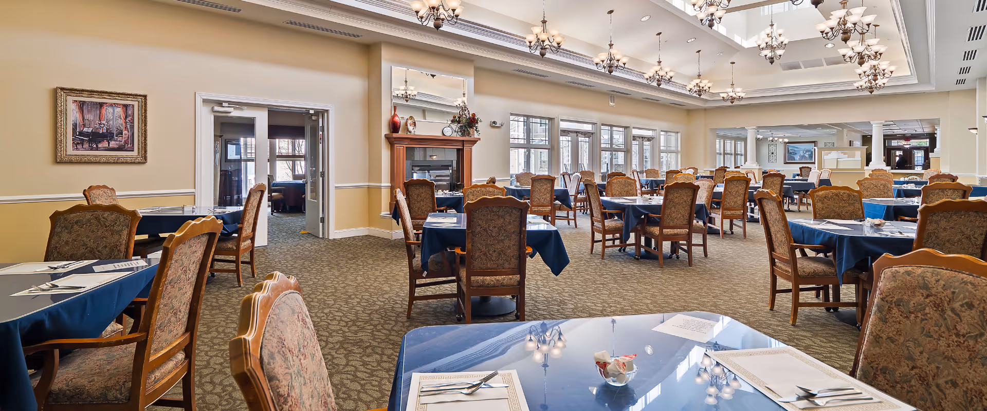 A spacious dining room with multiple tables covered in blue tablecloths and set with menus, napkins, and silverware. The room features patterned upholstered chairs, large windows letting in natural light, chandeliers hanging from a high ceiling with skylights, and a fireplace with decorative items on the mantel. The walls are painted beige with white trim, and there is a framed painting on one wall.