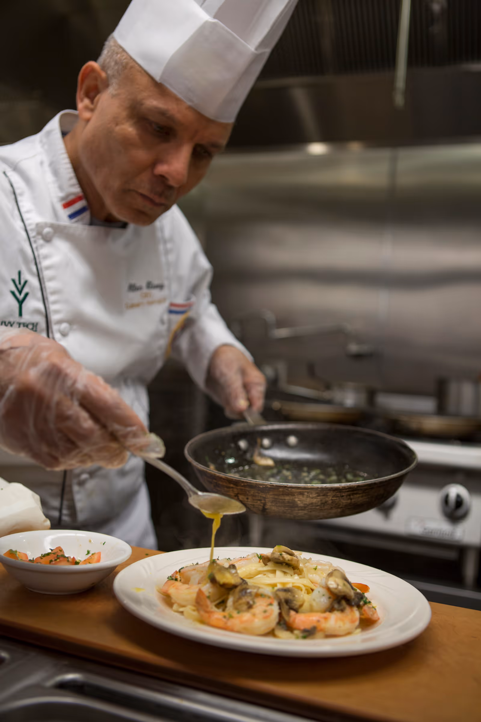 A chef wearing a white uniform and hat carefully pours sauce from a spoon onto a plated dish of pasta with shrimp and mushrooms in a professional kitchen.