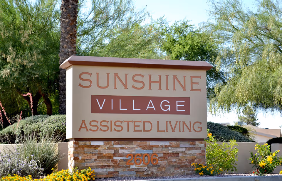 Outdoor sign for Sunshine Village Assisted Living with the address number 2606, surrounded by greenery and flowers under a clear sky.