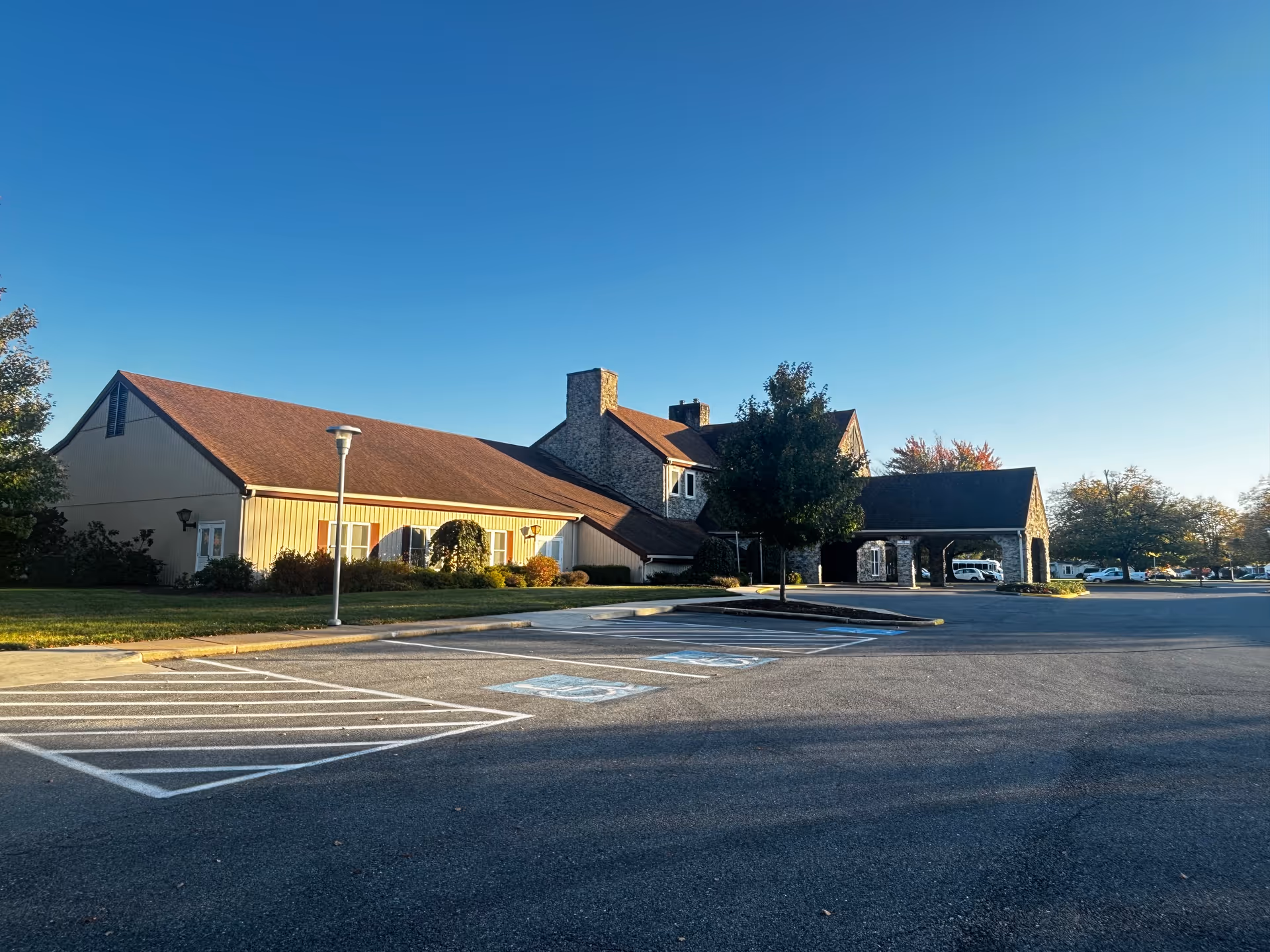 Exterior view of a senior living facility building with a stone and beige siding facade, a brown roof, and a covered entrance. The foreground shows an empty parking lot with marked handicap parking spaces and a clear blue sky above.