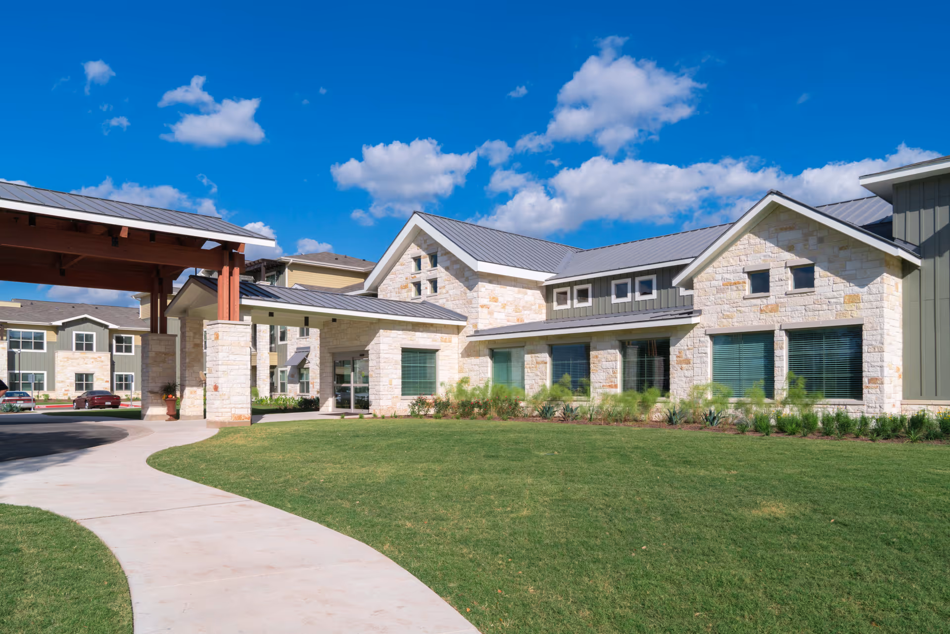 Exterior view of The Delaney at Georgetown Village senior living facility showing a modern building with stone and siding facade, large windows, a covered entrance, a curved sidewalk, and a well-maintained lawn under a blue sky with scattered clouds.