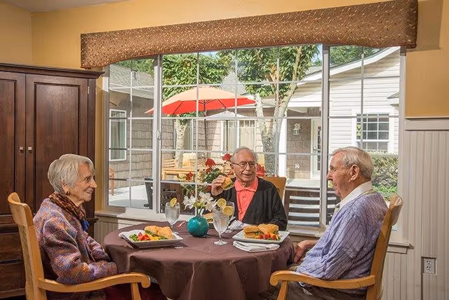 Three elderly people sitting around a round dining table with a brown tablecloth, enjoying a meal together in a room with large windows showing an outdoor patio with trees and a red umbrella.