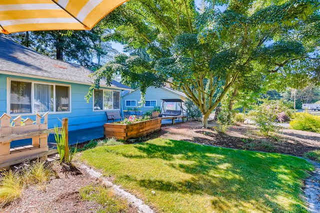A sunny outdoor garden area next to a light blue building with large windows. The garden features a wooden raised planter box with flowers, a bench, a large tree providing shade, and well-maintained grass and landscaping.