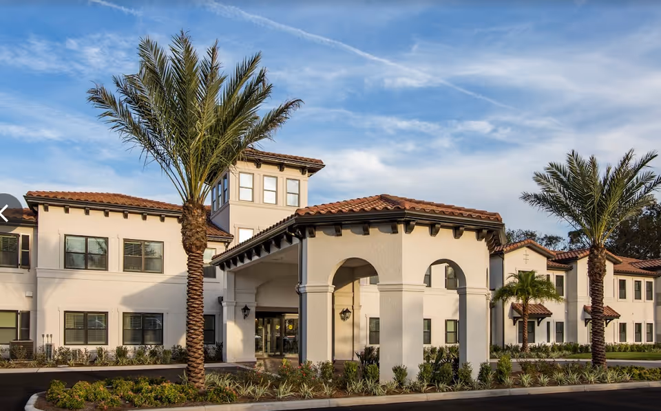 Front exterior of a Mediterranean-style senior living building with an arched entrance, red tile roof and palm trees.