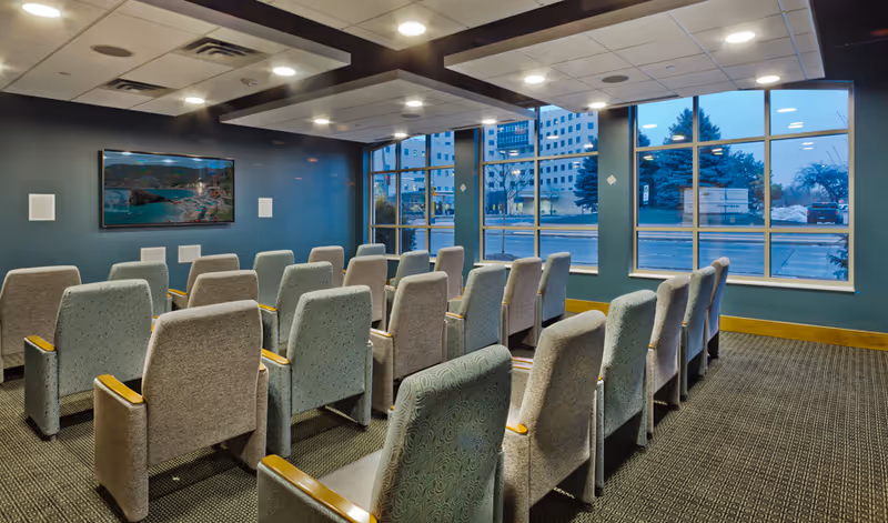 A small theater-style room with several rows of cushioned chairs facing a large flat-screen TV mounted on a blue wall. The room has large windows on one side showing an outside view of buildings and trees. The ceiling has recessed lighting and ventilation panels.