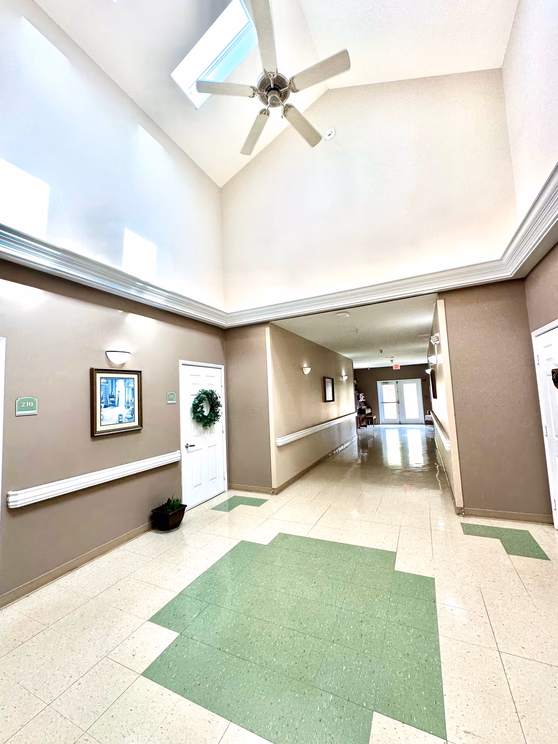 Bright interior hallway of an assisted living facility with a vaulted ceiling, ceiling fan and skylight, doors with room numbers, and green patterned tile flooring.
