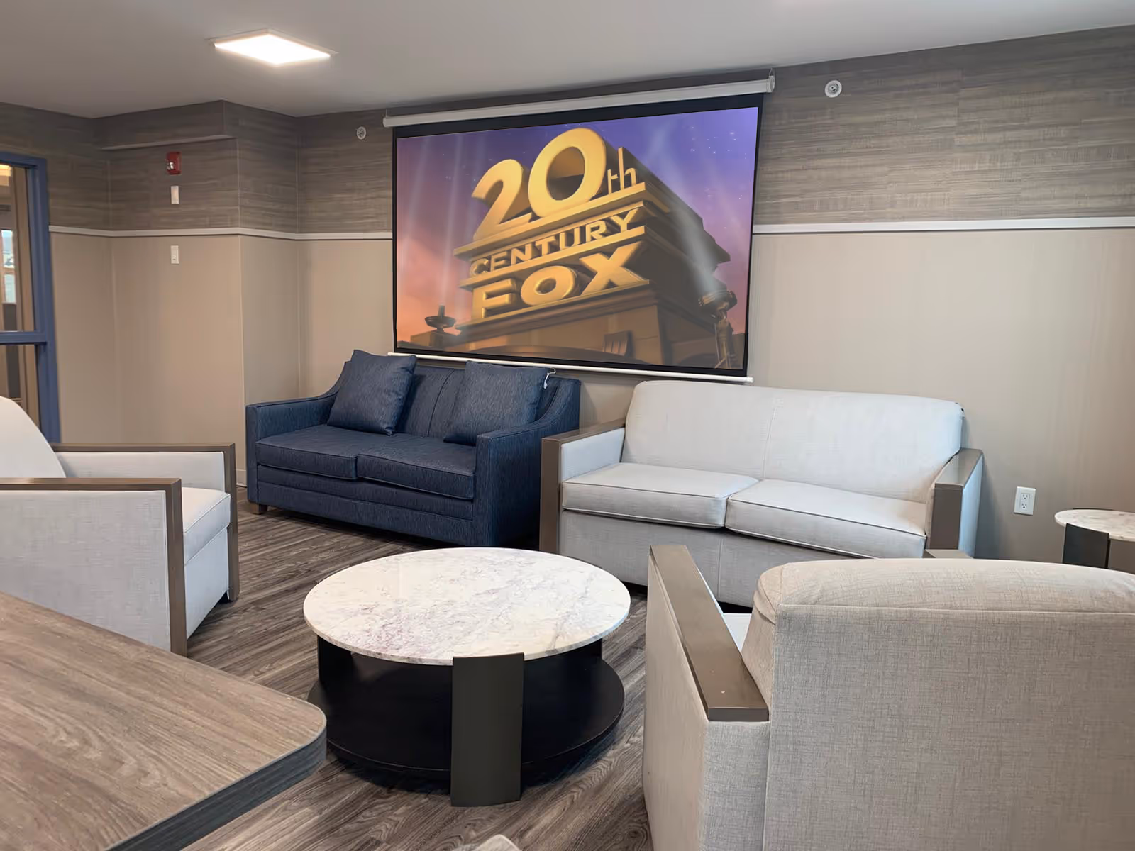 A cozy lounge area with two sofas, two armchairs, and a round marble coffee table in the center. A large screen on the wall displays the 20th Century Fox logo. The room has wood flooring and neutral-toned walls with a modern, comfortable design.