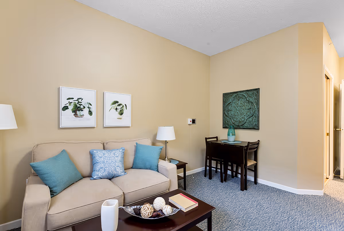 A cozy living room area with a beige sofa adorned with three blue cushions. Above the sofa are two framed pictures of green plants. To the right, there is a small dark wooden dining table with two chairs and a decorative green wall hanging above it. The room has beige walls and blue carpeted flooring. A floor lamp is positioned next to the sofa, and a coffee table in front holds decorative items and a book.