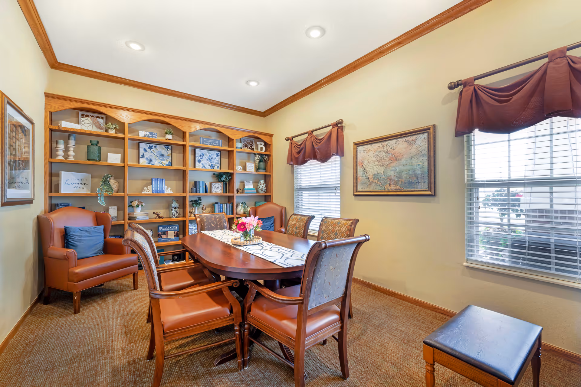 A warm dining room with a wooden table and upholstered chairs, built-in bookshelves, and windows with blinds.