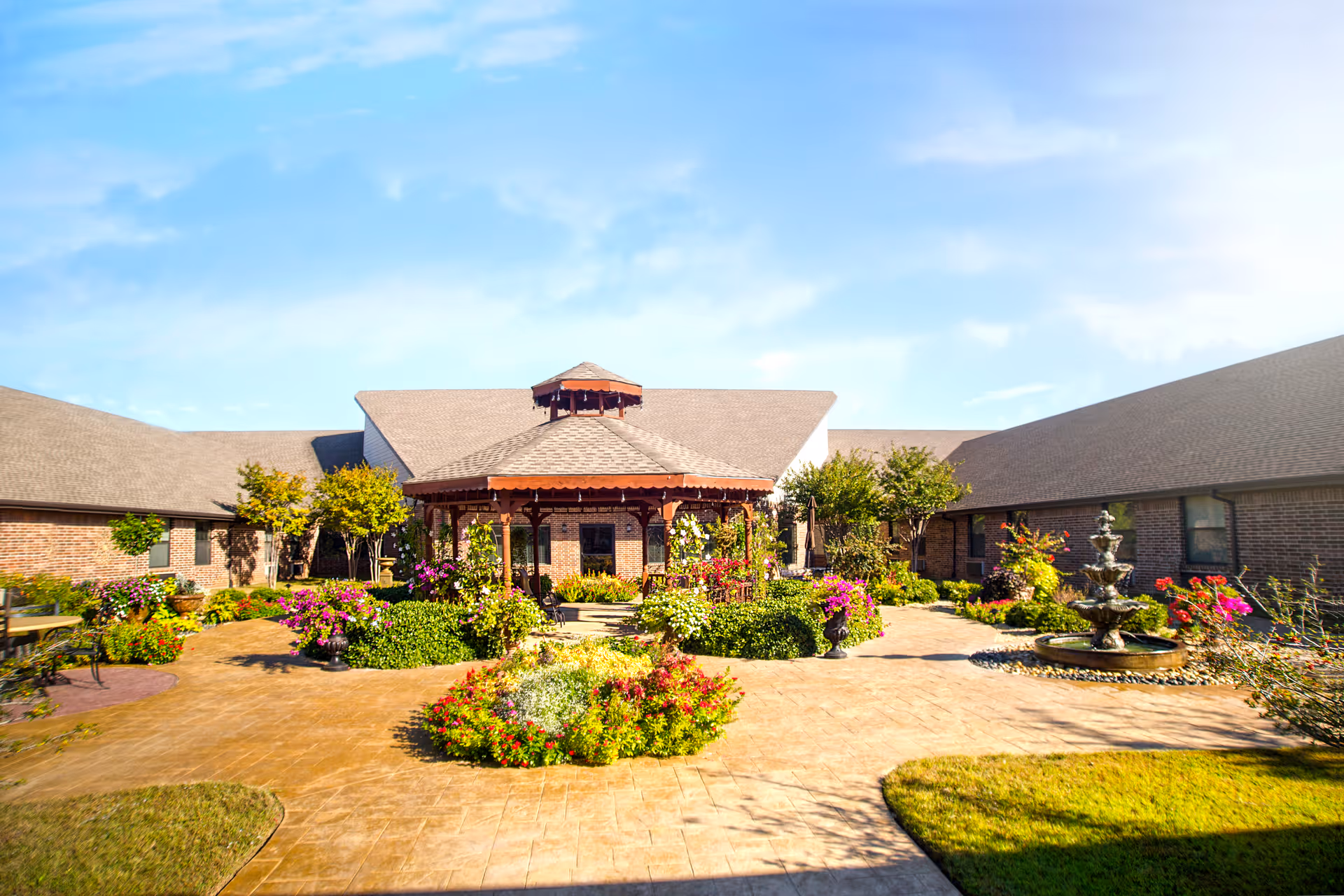 A bright outdoor courtyard with a central gazebo surrounded by colorful flowers and greenery. The courtyard is paved with stone tiles and features a multi-tiered water fountain on the right side. The surrounding buildings have brick walls and sloped roofs under a clear blue sky.