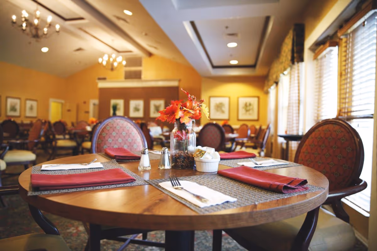 A warmly lit dining room with round wooden tables set with red napkins, silverware, salt and pepper shakers, and a small centerpiece of autumn leaves in a glass jar. The room has upholstered chairs, framed botanical artwork on the walls, chandeliers, and large windows with blinds allowing natural light in.
