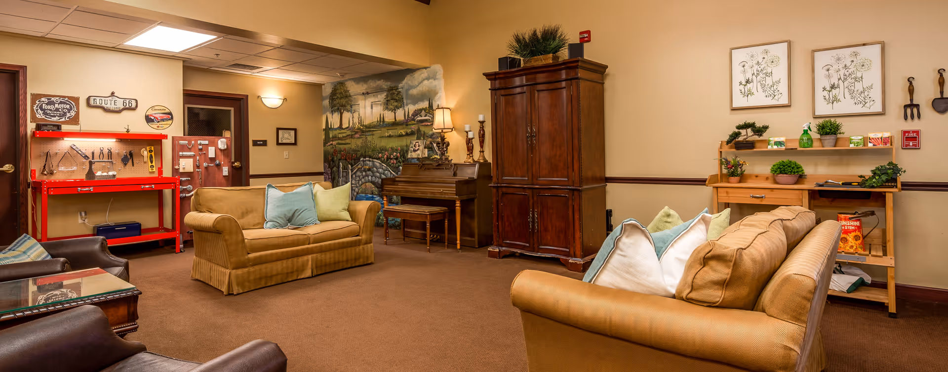 A cozy living room area in a senior living facility with two beige sofas adorned with pastel-colored pillows, a wooden piano against the wall, a large wooden cabinet, and a small wooden workbench with various tools and decorative plants. The walls are decorated with framed botanical prints, vintage signs, and a mural depicting a garden scene with trees, flowers, and a stone bridge.