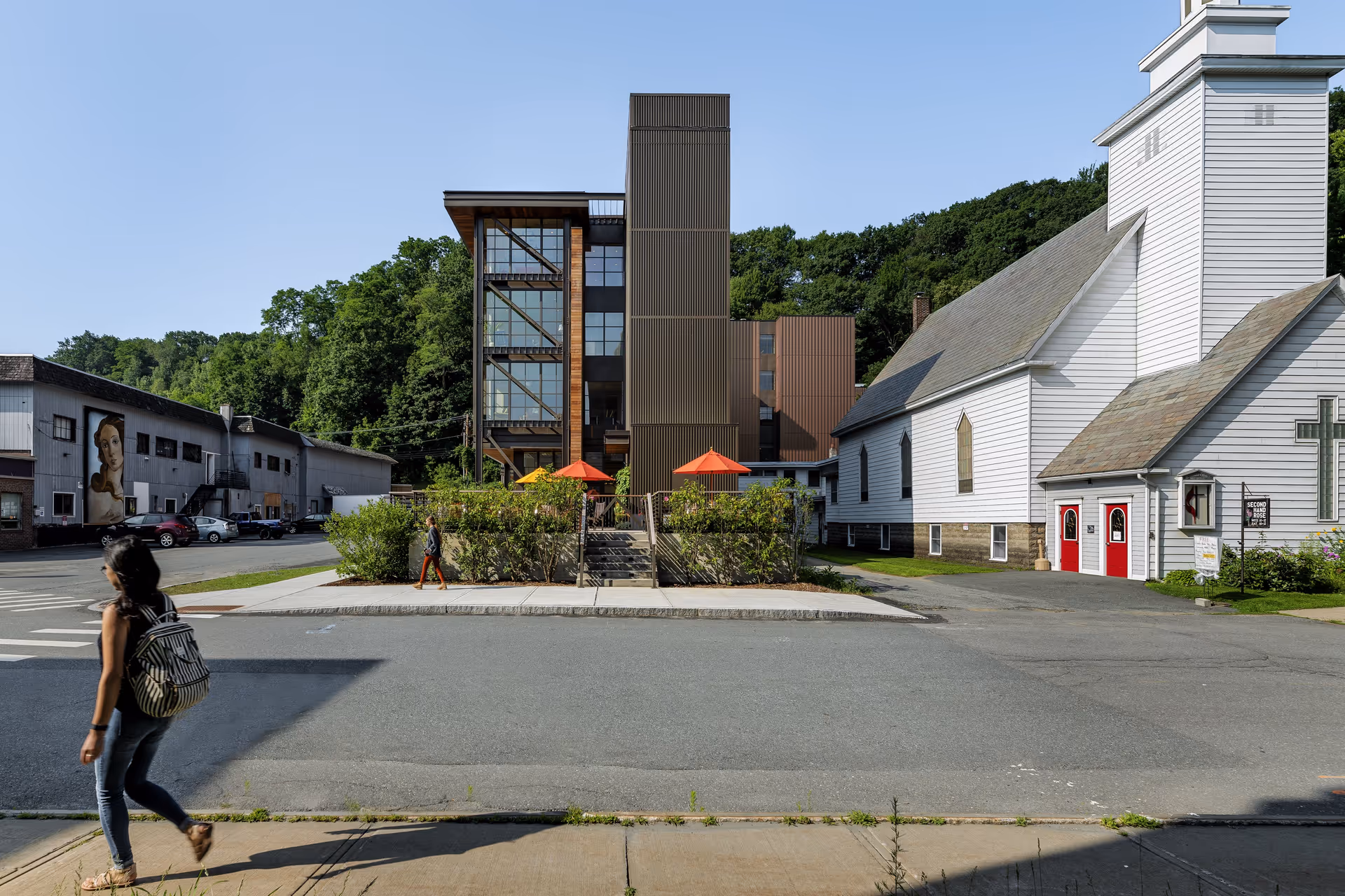 Street view showing a modern multi-story building with large glass windows and brown paneling, adjacent to a white church with red doors and a cross on the side. Two people are walking on the sidewalk and street in front of the buildings. Trees and greenery are visible in the background under a clear blue sky.