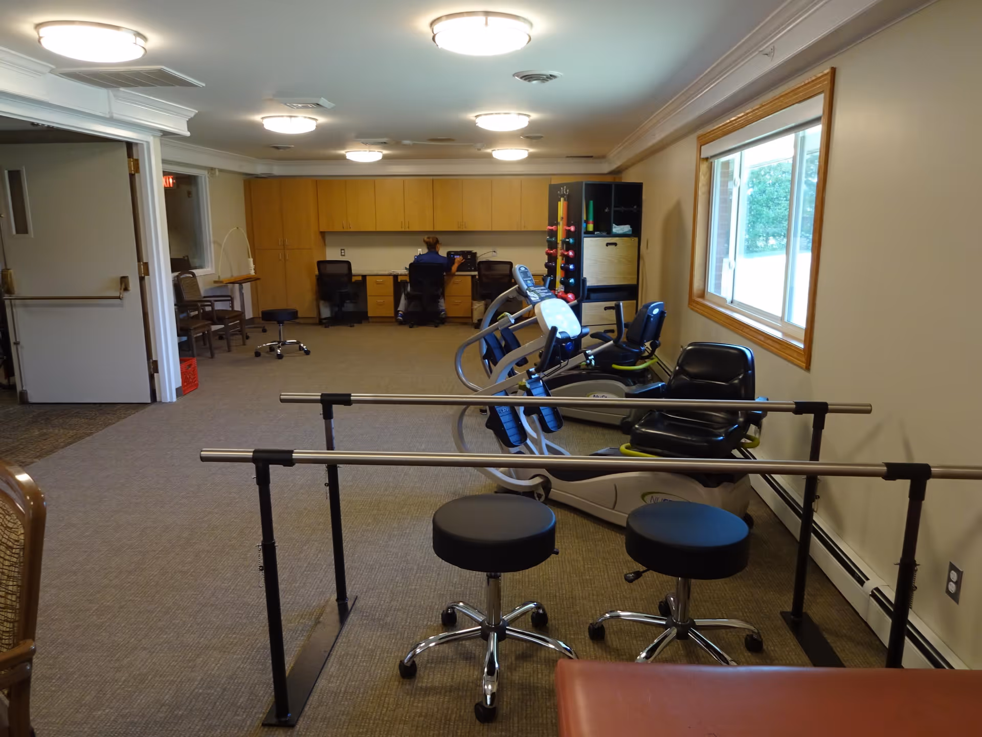 Interior therapy/fitness room with parallel bars, exercise bikes, stools and a desk area near a window.