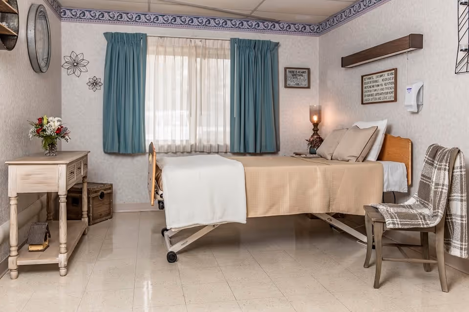 A neatly arranged nursing facility bedroom with a single bed covered in beige and white bedding. The room features a wooden chair with a plaid blanket draped over it, a small wooden table with a flower vase, and two windows with blue curtains and sheer white drapes. The walls are decorated with framed inspirational quotes and metal flower wall art. The floor is tiled and the room has a calm, homey atmosphere.