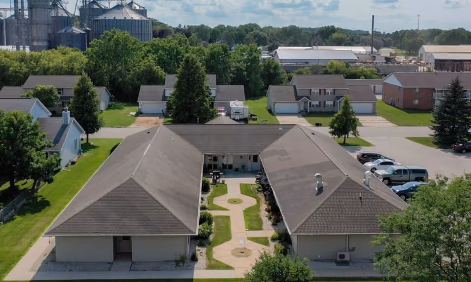 Aerial view of Scandinavian Court Assisted Living facility showing a U-shaped building with a central courtyard featuring a paved walkway and greenery. Surrounding the facility are residential houses, trees, parked cars, and industrial buildings in the background under a partly cloudy sky.