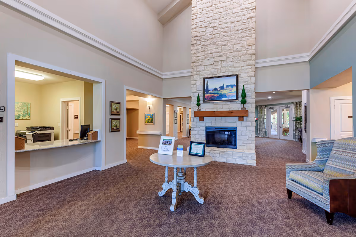 Interior view of a senior living facility lobby with a high ceiling and a stone fireplace. A round wooden table with informational materials is centered on a carpeted floor. To the right is a patterned armchair, and to the left is a reception window with office equipment visible inside. The walls are decorated with framed artwork and the entrance doors are visible in the background.