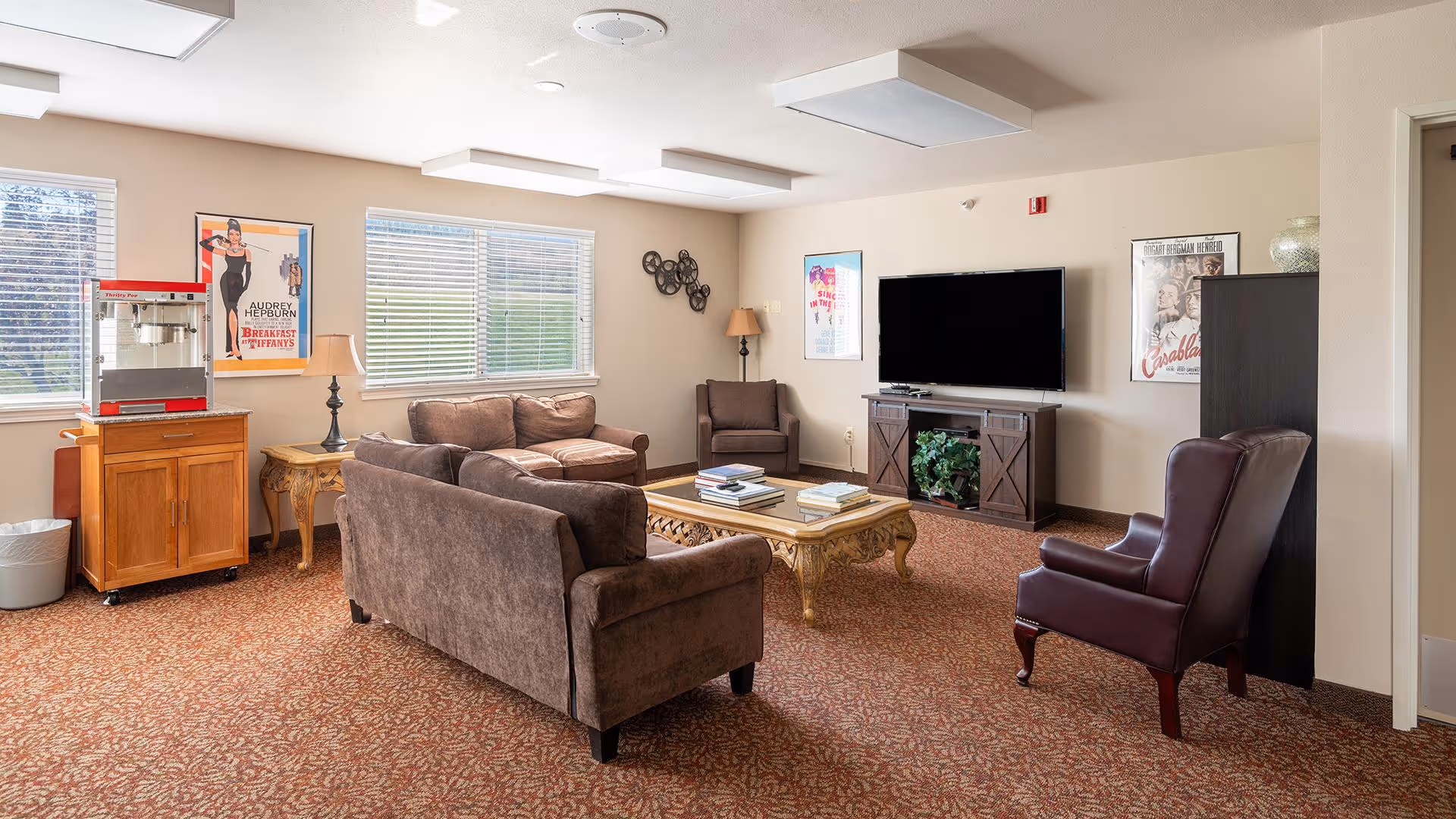 A cozy senior living facility common room with brown upholstered sofas and armchairs arranged around a wooden coffee table. A large flat-screen TV is mounted on a wooden cabinet against the wall. The room has beige walls with framed vintage movie posters and two windows with blinds letting in natural light. A popcorn machine sits on a wooden cart near the windows, and the floor is covered with patterned carpet.