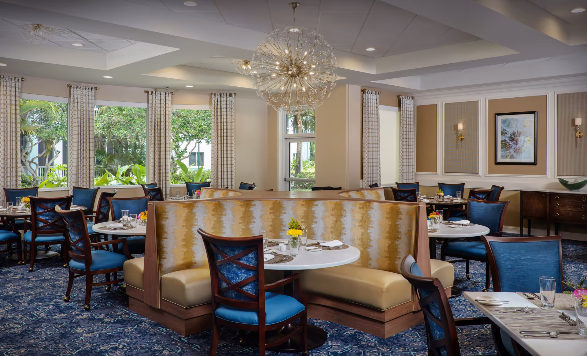 A bright and elegant dining room with round tables set with plates, glasses, and silverware. The room features blue upholstered chairs and a curved booth with a yellow and beige patterned backrest. Large windows with patterned curtains allow natural light to fill the space, and a modern chandelier hangs from the ceiling. The walls are decorated with framed artwork and sconces.