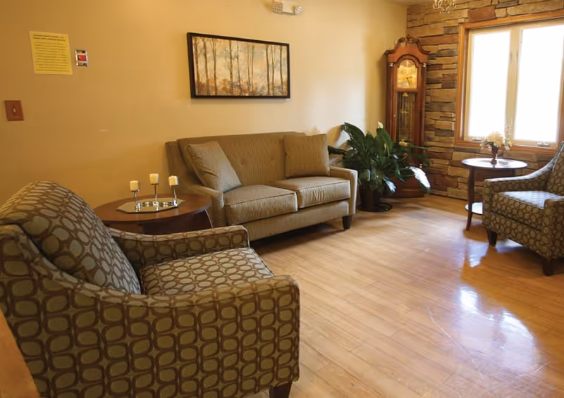 Comfortable sitting room with a sofa, patterned armchairs, side tables, a potted plant and a grandfather clock beside a window.