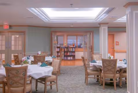 Senior living dining room with round tables, chairs, white tablecloths, and a library in back.