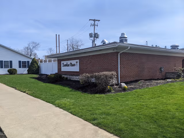 Exterior view of a single-story brick building with a sign that reads 'Brookline Manor'. The building is surrounded by green grass, some bushes, and a concrete sidewalk. Another white building is visible in the background under a clear blue sky.