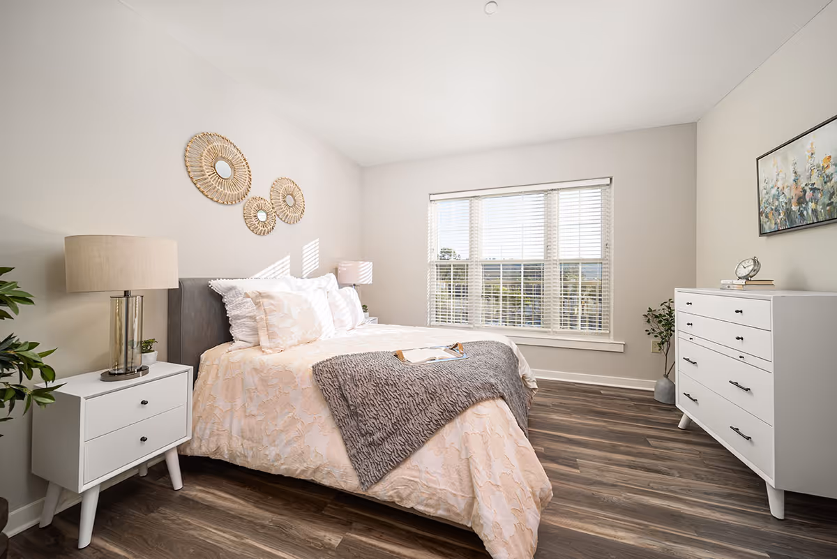 A bright and airy bedroom with a large window covered by white blinds. The room features a bed with a light pink floral bedspread, white pillows, and a gray throw blanket. There are two white nightstands with lamps on either side of the bed, a white dresser with books and a clock on top, and decorative round mirrors on the wall above the bed. The floor is wood with a dark finish.