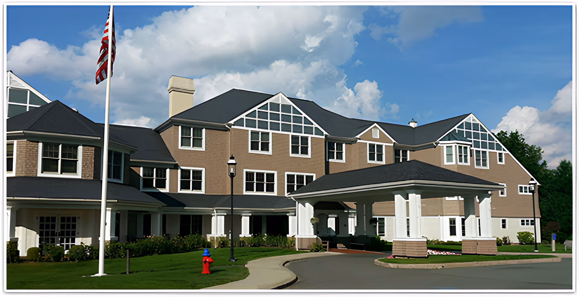Front exterior of a multi-story senior living building with a covered entrance, flagpole, and landscaped lawn.