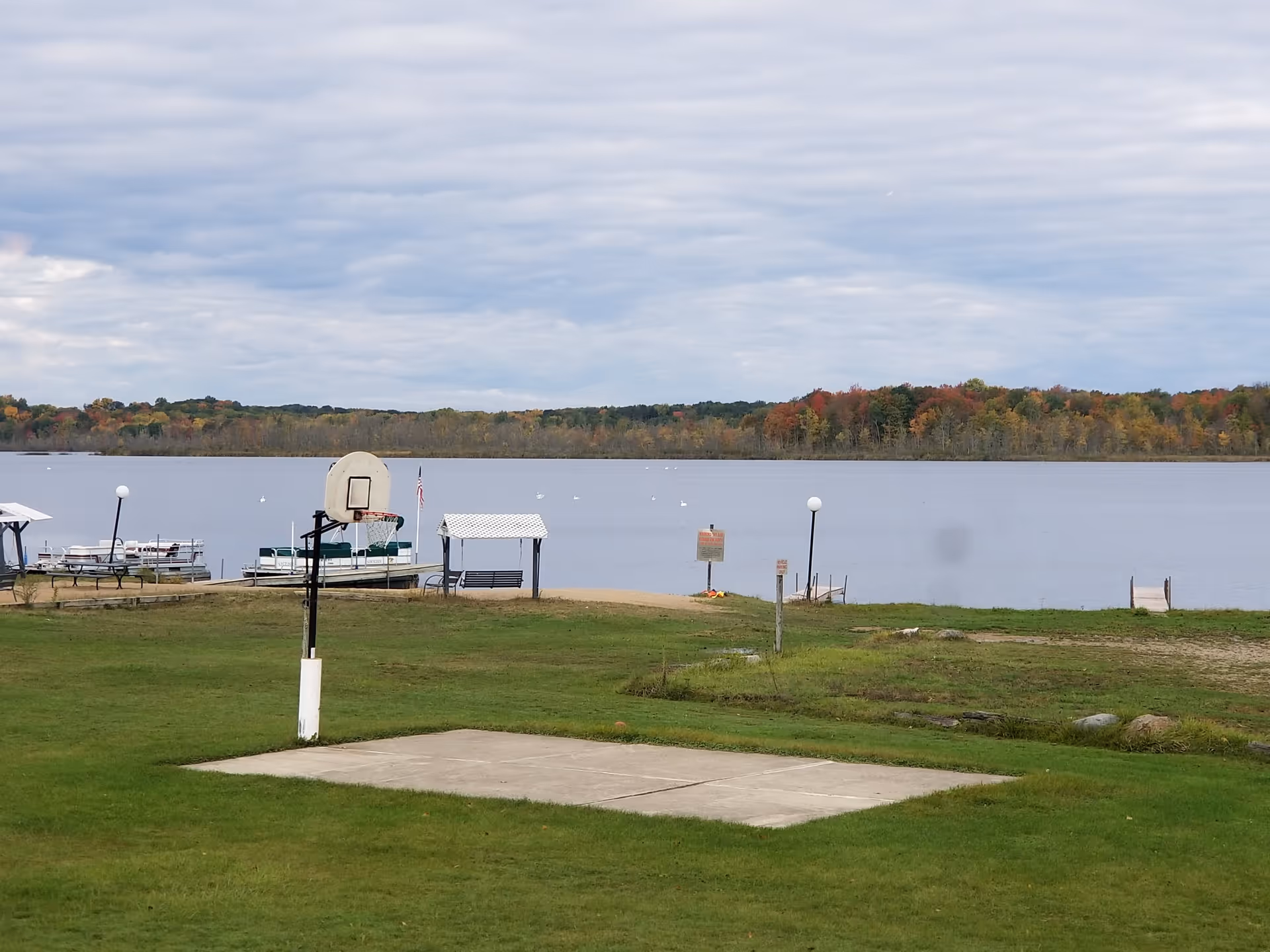 Outdoor scene at Lakeside Village 55+ Waterfront Community showing a small concrete basketball court with a single hoop, green grassy area, a lake with docks and boats, and a tree line with autumn foliage under a cloudy sky.