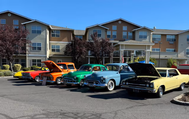 Several colorful classic cars with some hoods open parked in front of a multi-story senior living building under a clear blue sky.
