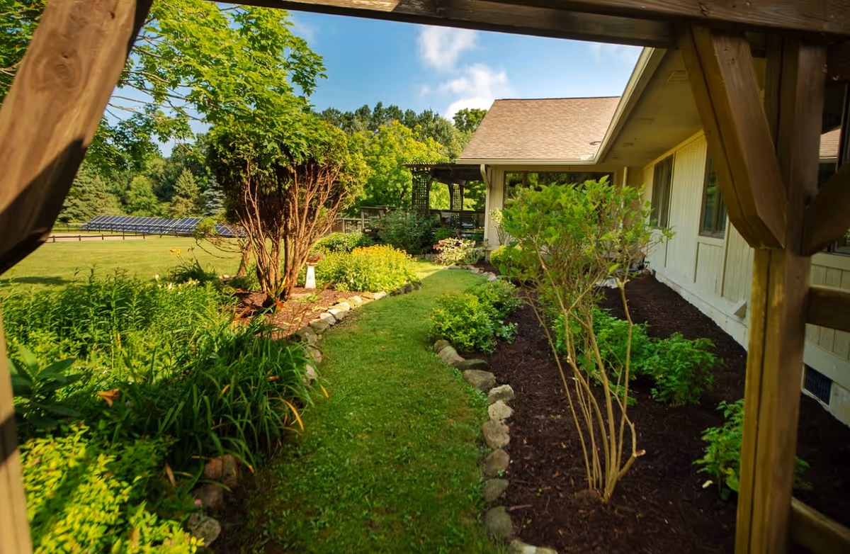A grassy garden path lined with flowerbeds and shrubs running alongside a single-story building framed by a wooden pergola.
