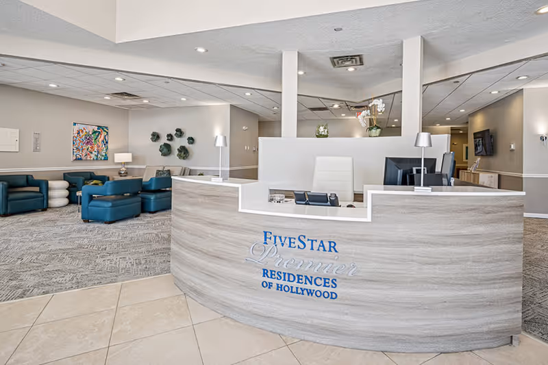 Reception area of Five Star Premier Residences of Hollywood featuring a curved wooden reception desk with a white countertop, two desk lamps, computer monitors, and a white office chair. Behind the desk is a seating area with teal armchairs, a beige sofa, a side table with a lamp, and wall decorations including a colorful abstract painting and green wall art. The floor is tiled near the desk and carpeted in the seating area.