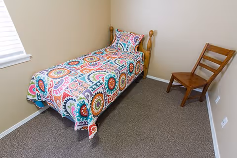 Small bedroom with a single wooden bed covered by a colorful patterned quilt, a wooden chair, beige walls, a window, and carpeted floor.