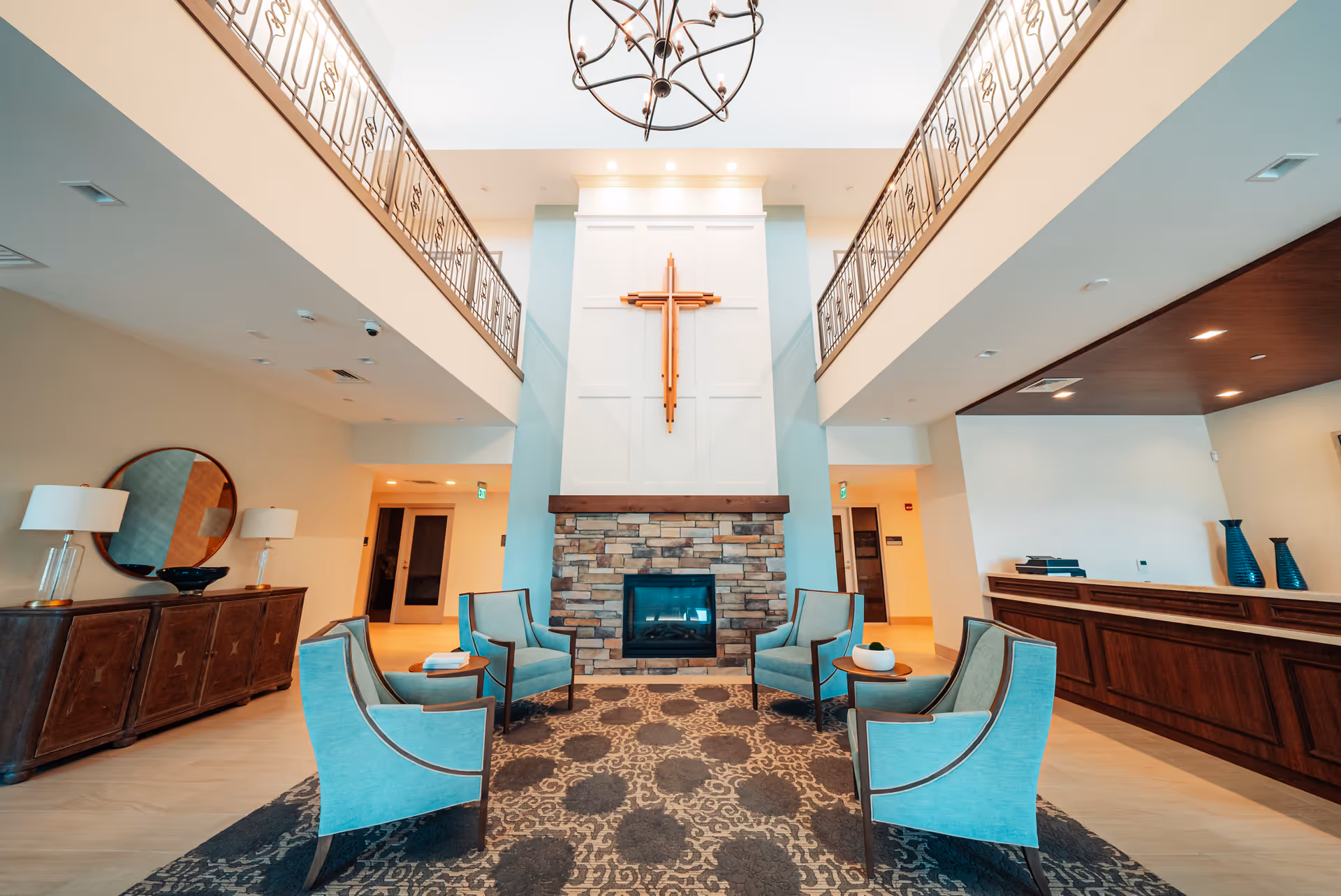 A spacious and well-lit common area with a high ceiling and a large wooden cross mounted above a stone fireplace. Four light blue armchairs are arranged around a small round table on a patterned carpet. To the left, there is a wooden sideboard with two lamps and a round mirror above it. To the right, there is a wooden reception desk with decorative vases. The upper level has a railing overlooking the area.