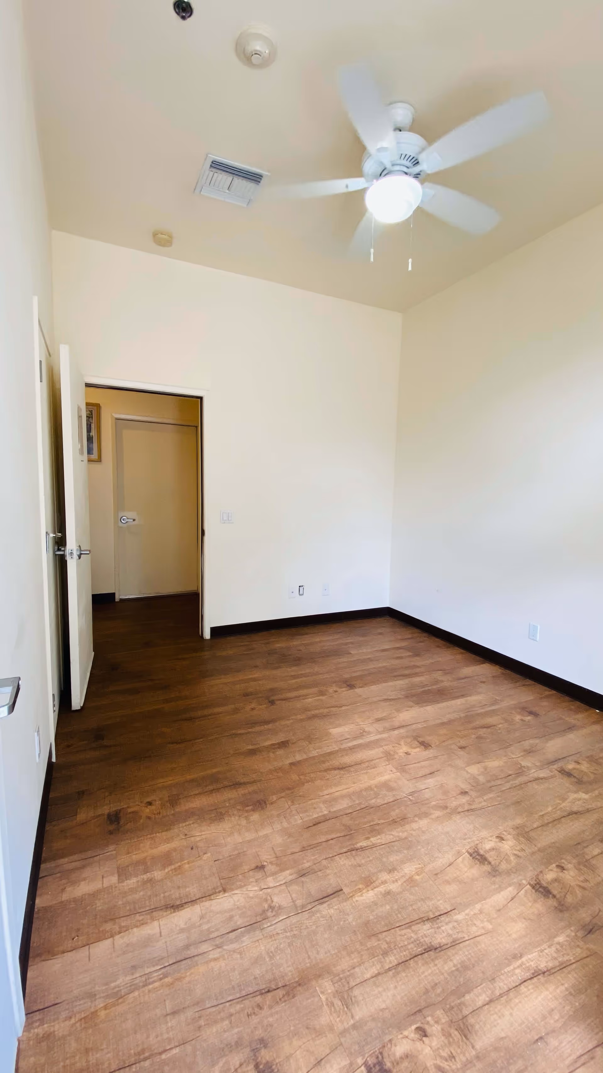 Empty room with wooden flooring, cream-colored walls, a white ceiling fan with light, and an open door leading to another room with a closed door.