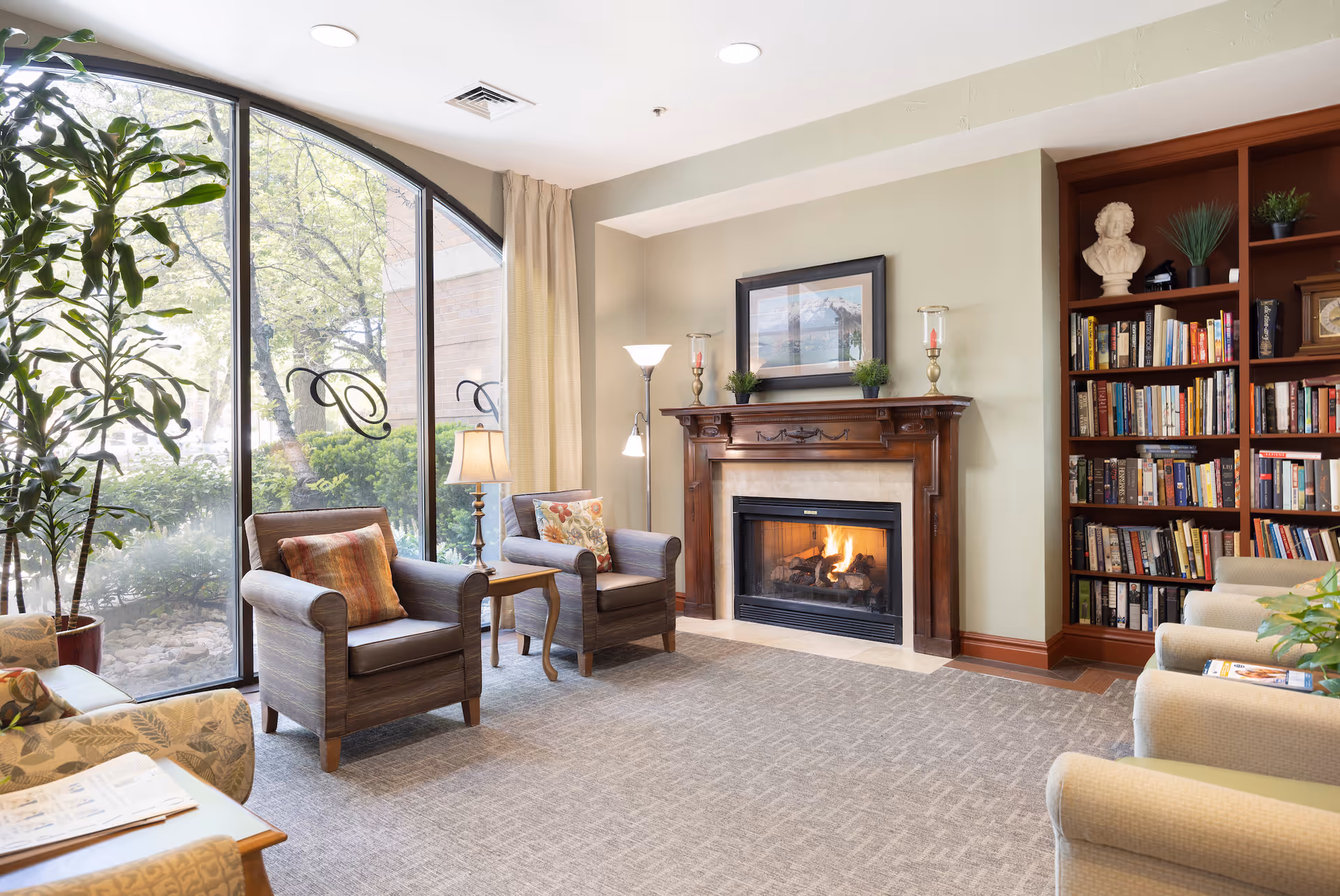 Bright senior living room lounge with a fireplace, armchairs, bookshelves and a large arched window.