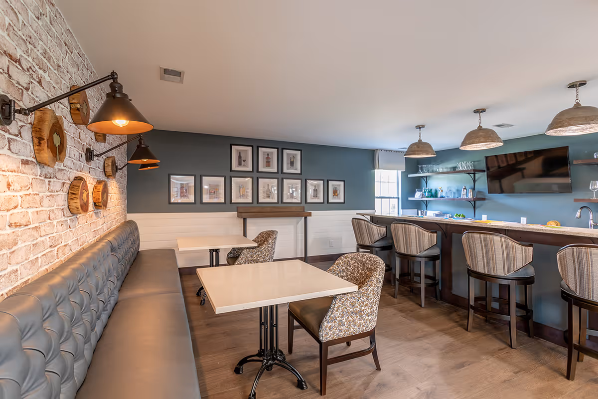 A cozy dining area with a long tufted gray bench along a brick wall, two small square tables with patterned chairs, and a bar counter with four cushioned bar stools. The wall behind the bar is painted blue and decorated with framed pictures and shelves holding glasses and bottles. Three pendant lights hang above the bar, and a flat-screen TV is mounted on the wall.