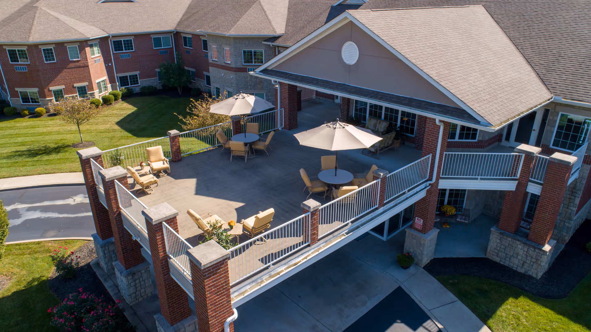 A spacious outdoor patio area at Magnolia Springs East Louisville with several round tables, chairs, and large umbrellas. The patio is elevated and supported by brick and stone pillars, overlooking a driveway and landscaped green lawn with bushes and small trees.