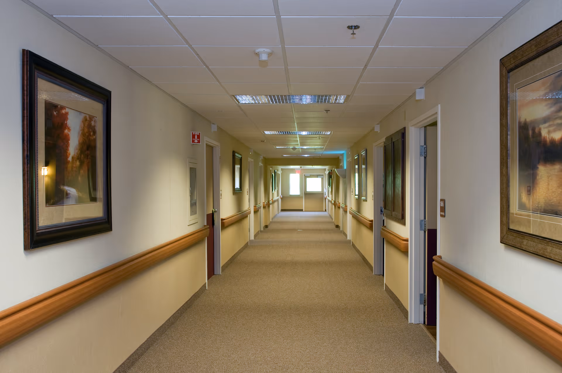Carpeted corridor in a senior living facility lined with wooden handrails, framed pictures, and multiple room doors.