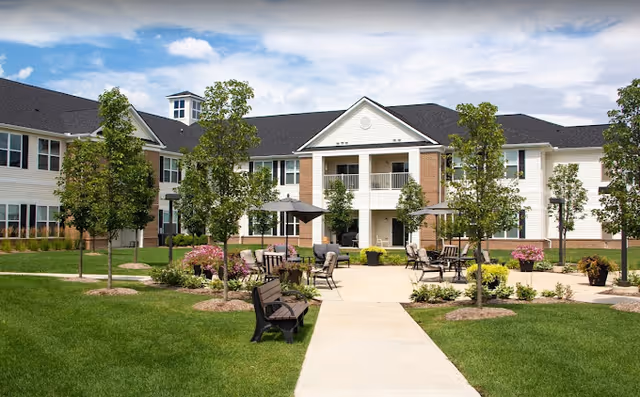Outdoor courtyard area of Olivia's Assisted Living facility featuring a paved walkway, benches, tables with umbrellas, trees, and landscaped flower beds with a two-story building in the background under a partly cloudy sky.